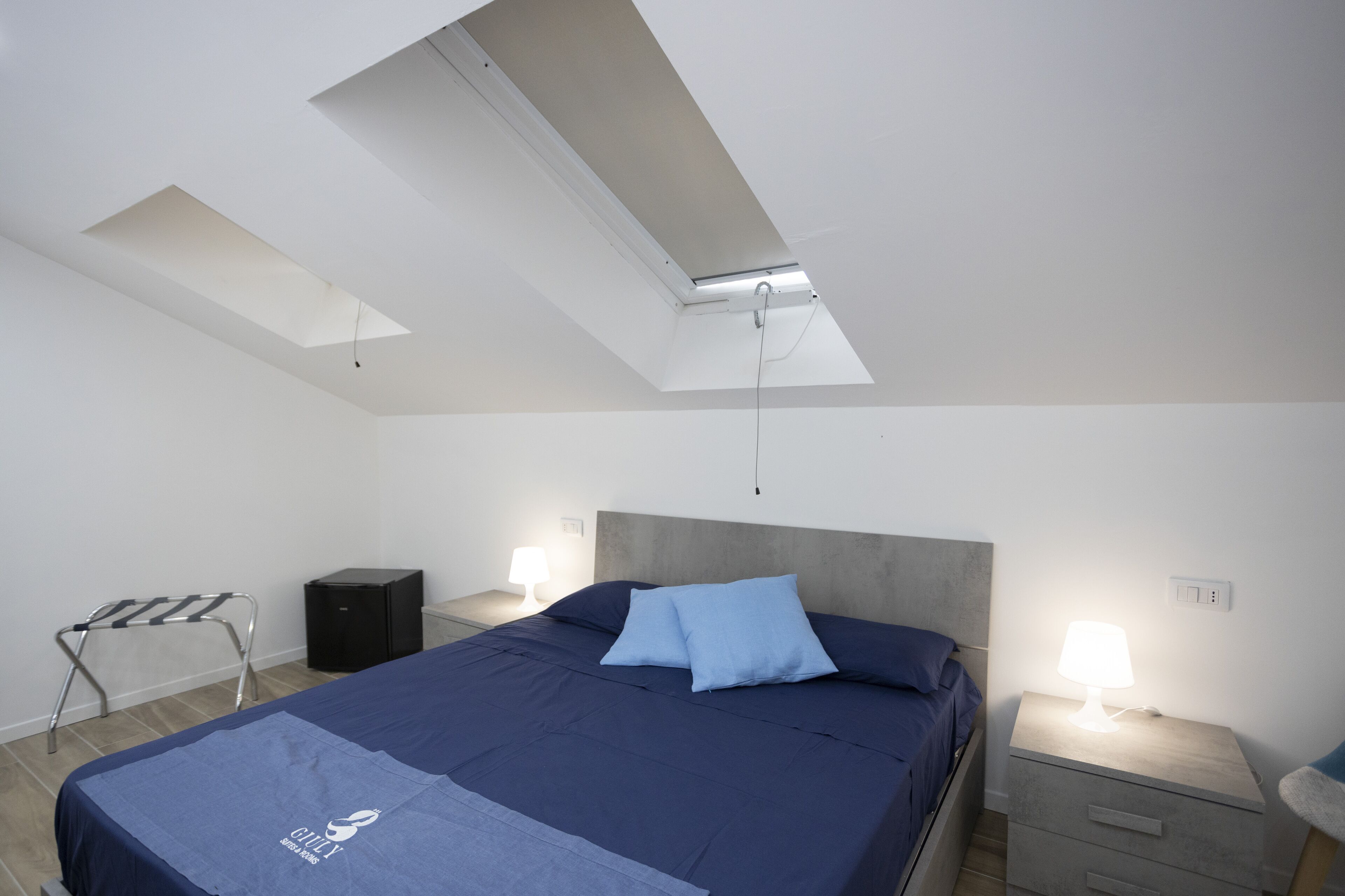 Ein modernes Schlafzimmer mit Dachfenster, einem Bett mit blauer Bettwäsche, einem Nachttisch mit Lampe und einem kleinen Schrank.