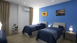 A hotel room with two beds, a chair, a nightstand with a lamp, a wall-mounted air conditioner, and a cityscape painting. - GIULY SUITES & ROOMS (Naples)