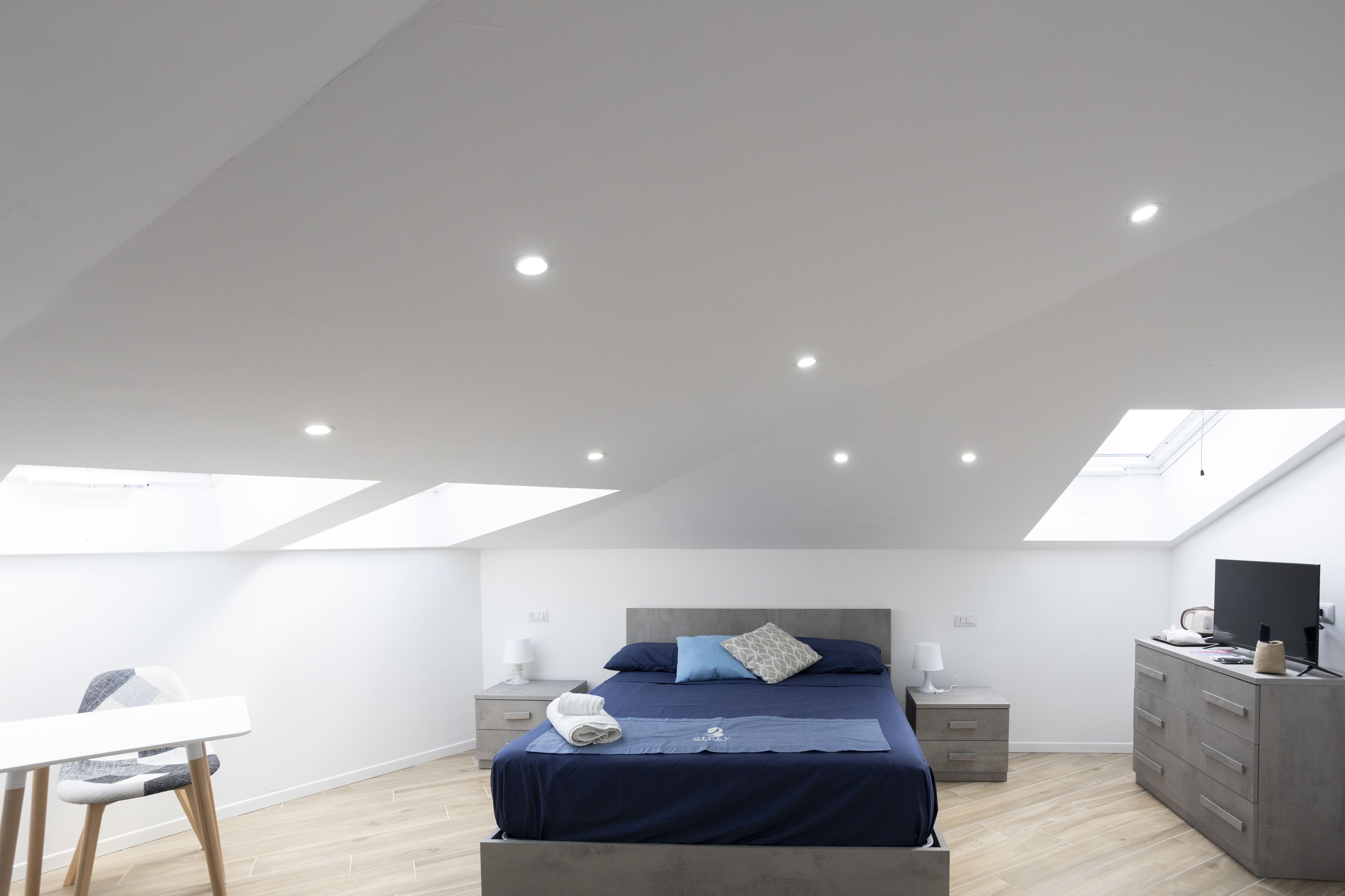 Un dormitorio moderno con tragaluz, una cama con colcha azul, una mesita de noche con lámpara y un aparador.