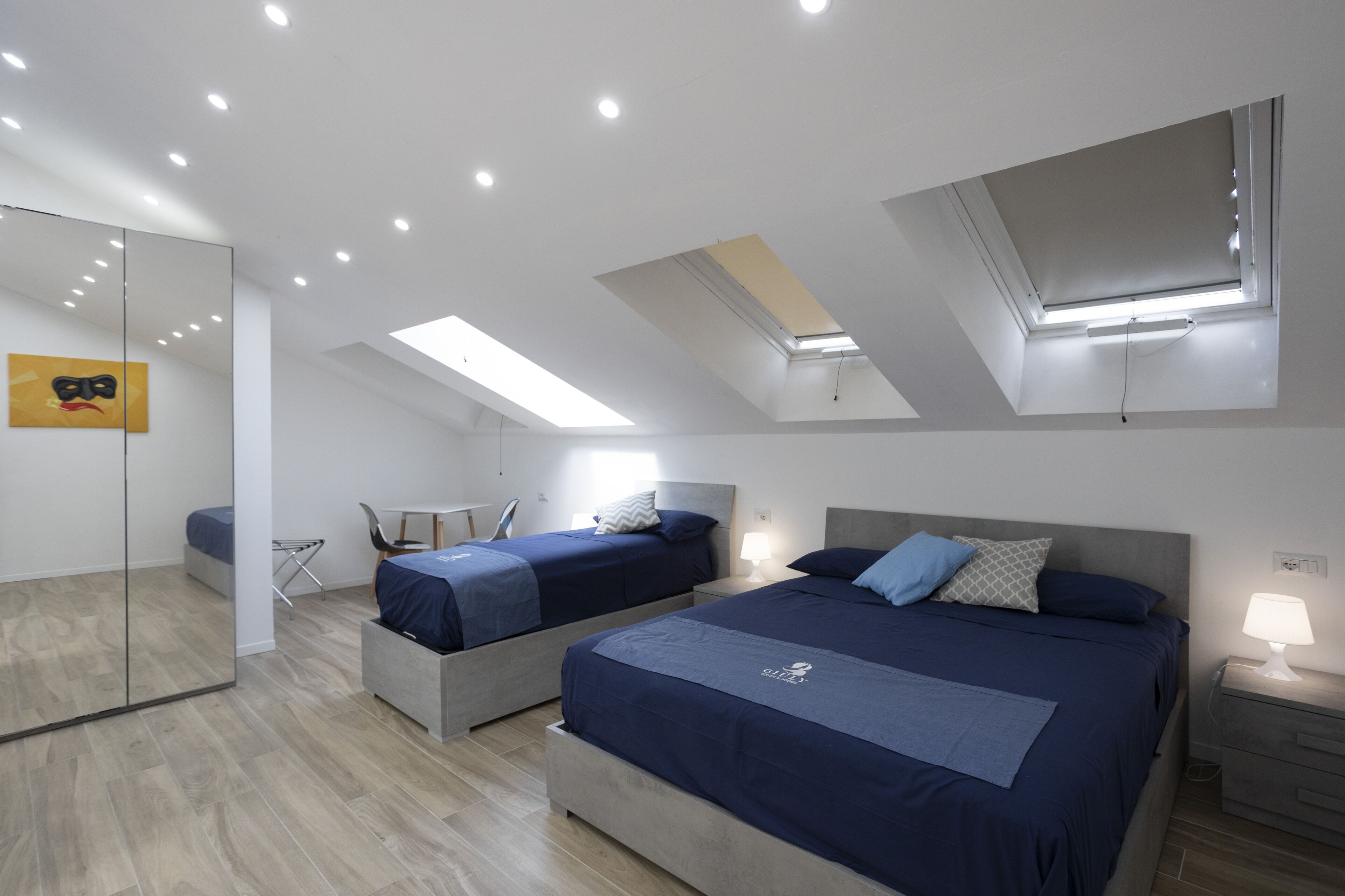 A modern bedroom with two beds, skylights, a mirror, and a bedside table with a lamp.