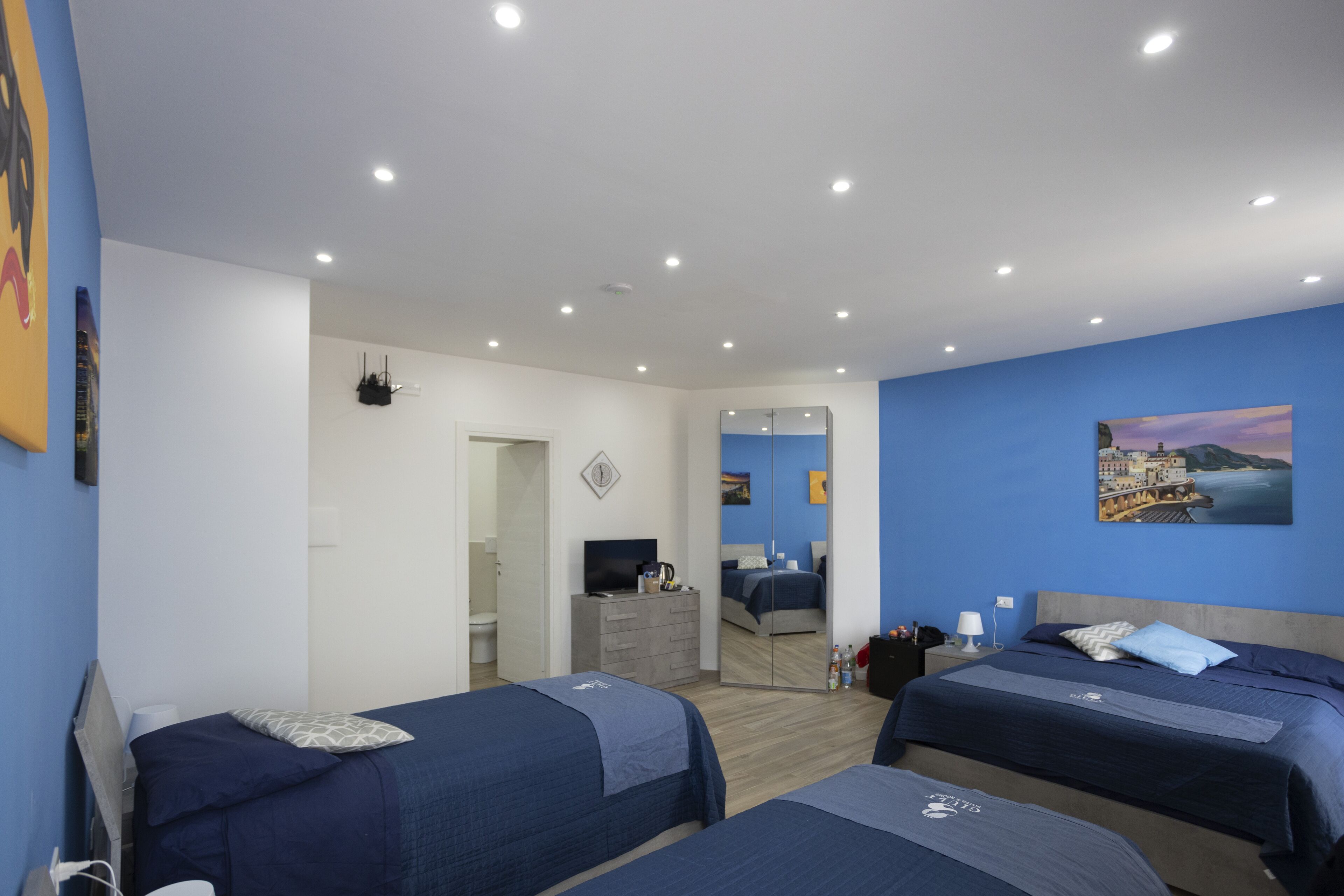 A modern hotel room with two single beds, a large mirror, a painting of a coastal scene, and a small desk with a TV.