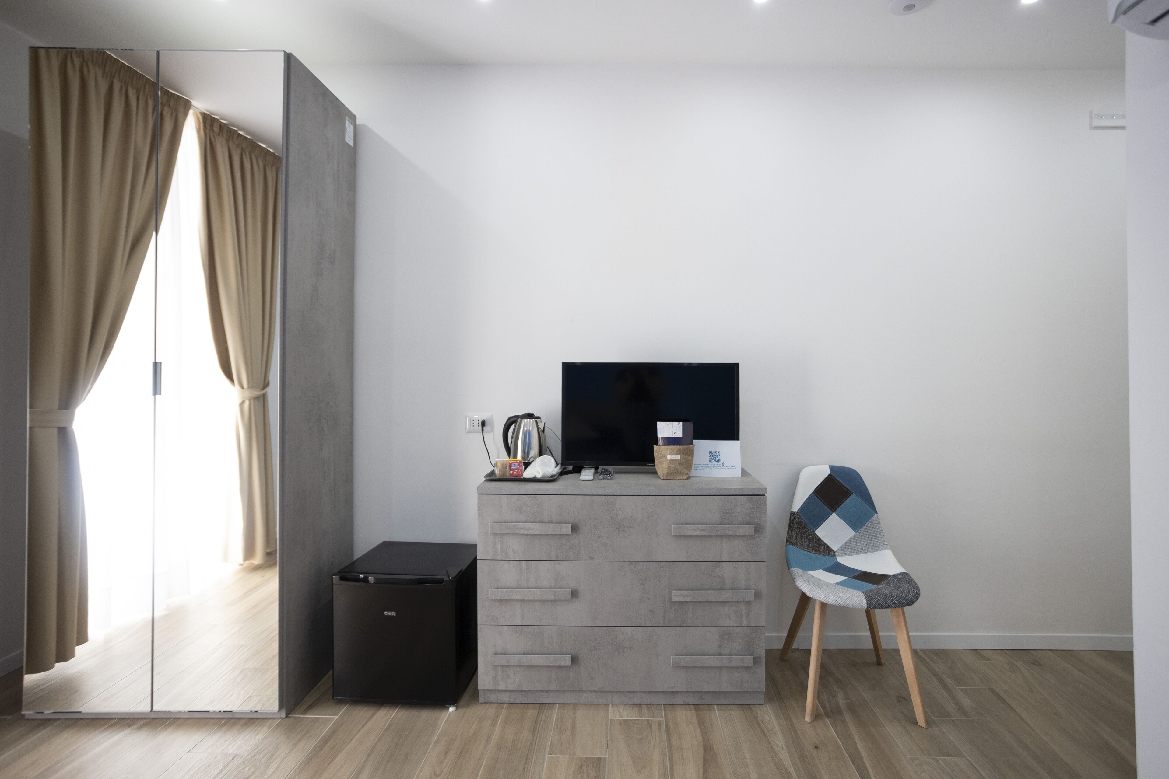 Habitació moderna amb un calaix gris, una televisió, un petit safataire i una cadira.