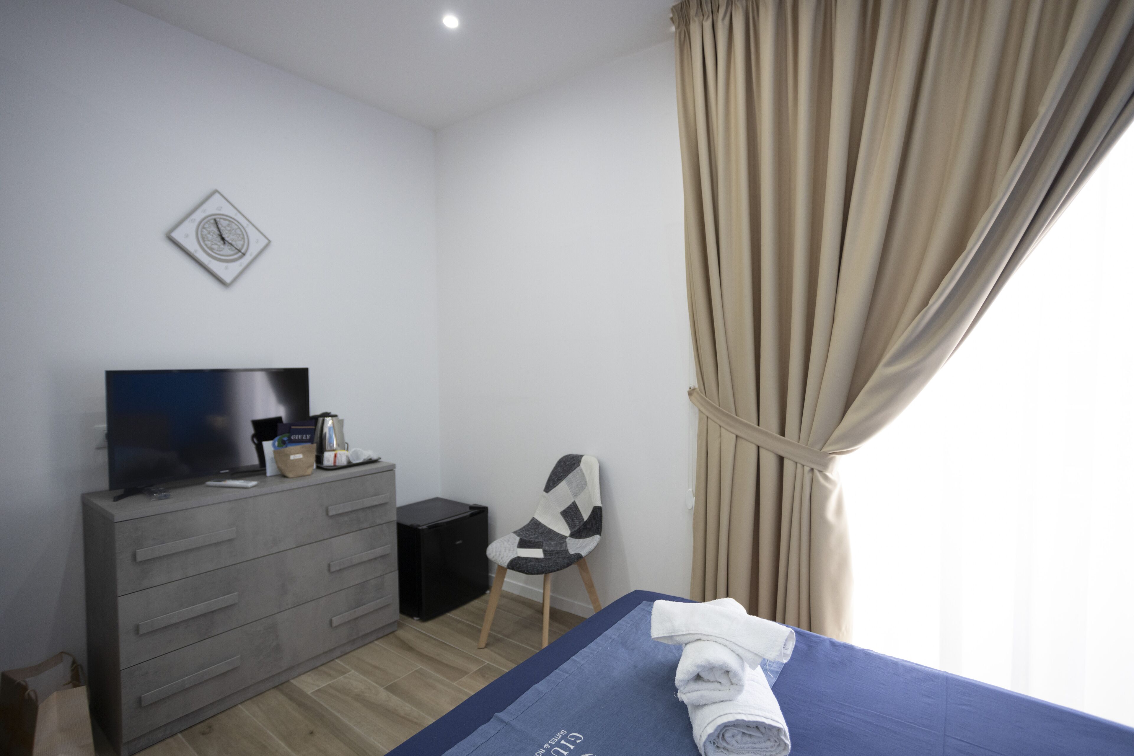 Une chambre d’hôtel avec un lit, une télévision, une commode, une chaise et une fenêtre avec des rideaux.