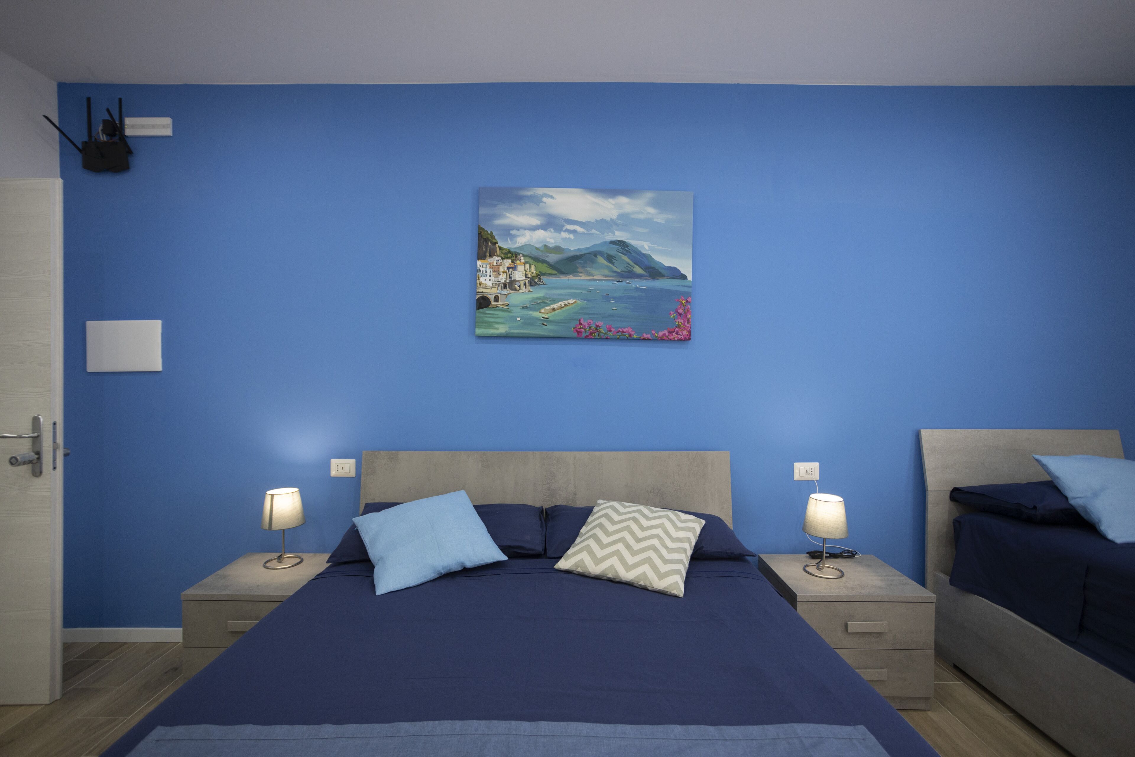 Un dormitorio con pared azul, una cama con cabecera de madera, dos mesitas de noche con lámparas y un cuadro que muestra una escena costera.