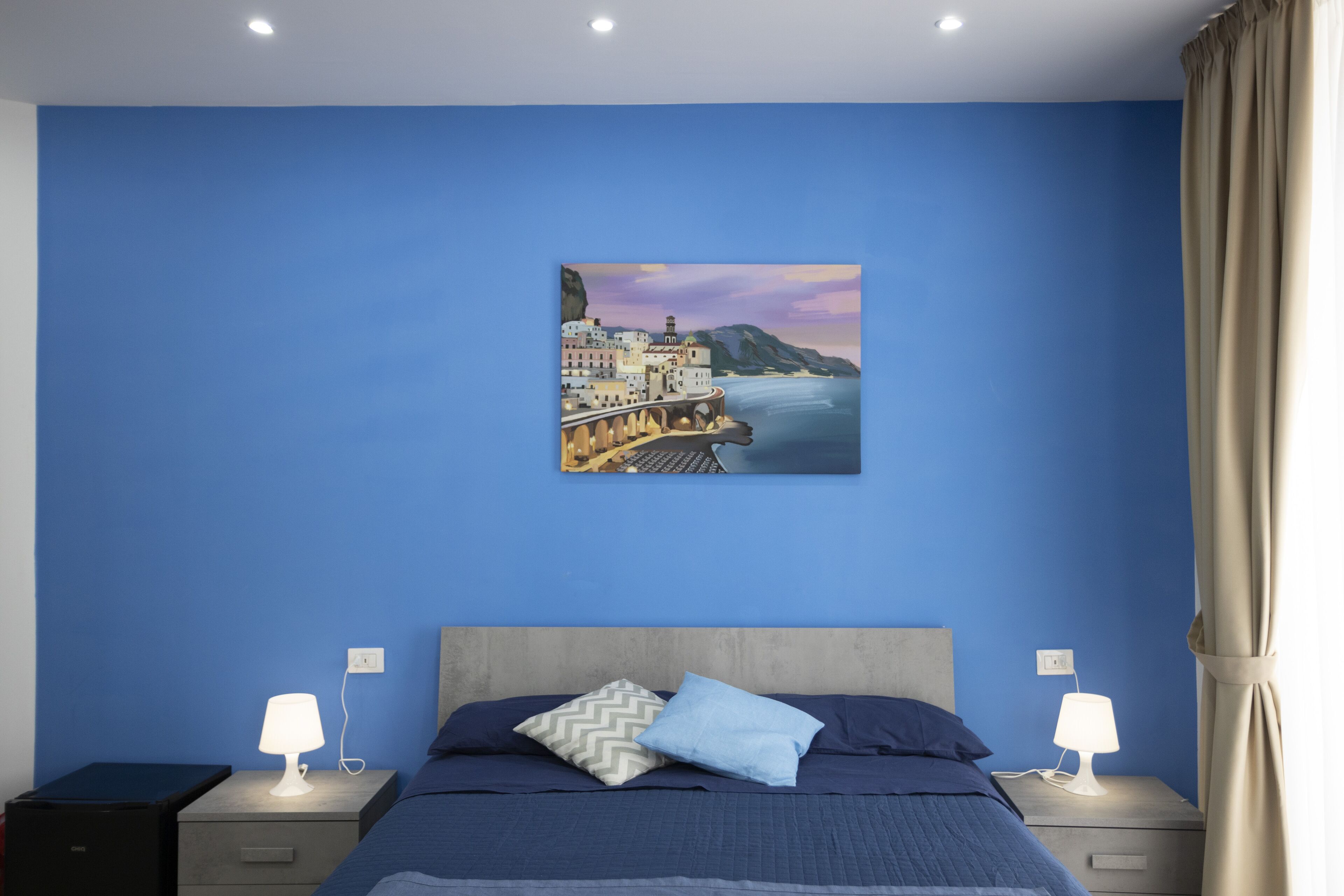 A bedroom with a bed, two bedside tables, a nightstand, and a wall-mounted painting of a coastal town.