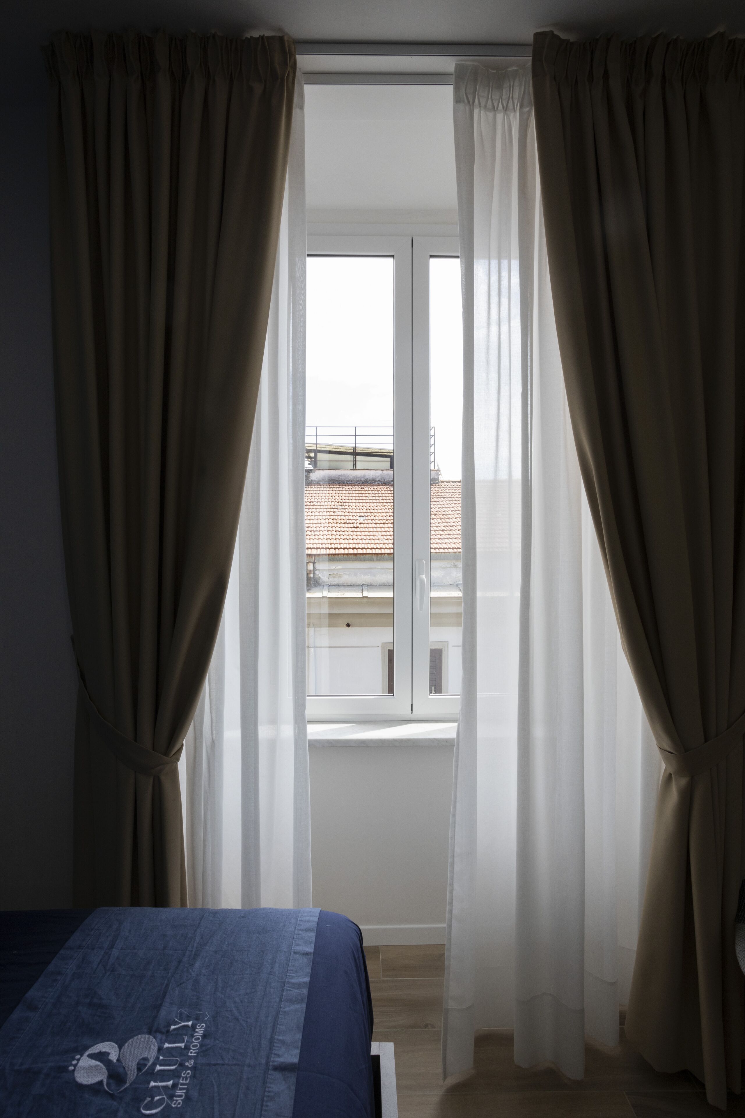 A bedroom with a bed, beige curtains, a window, and a view of buildings.