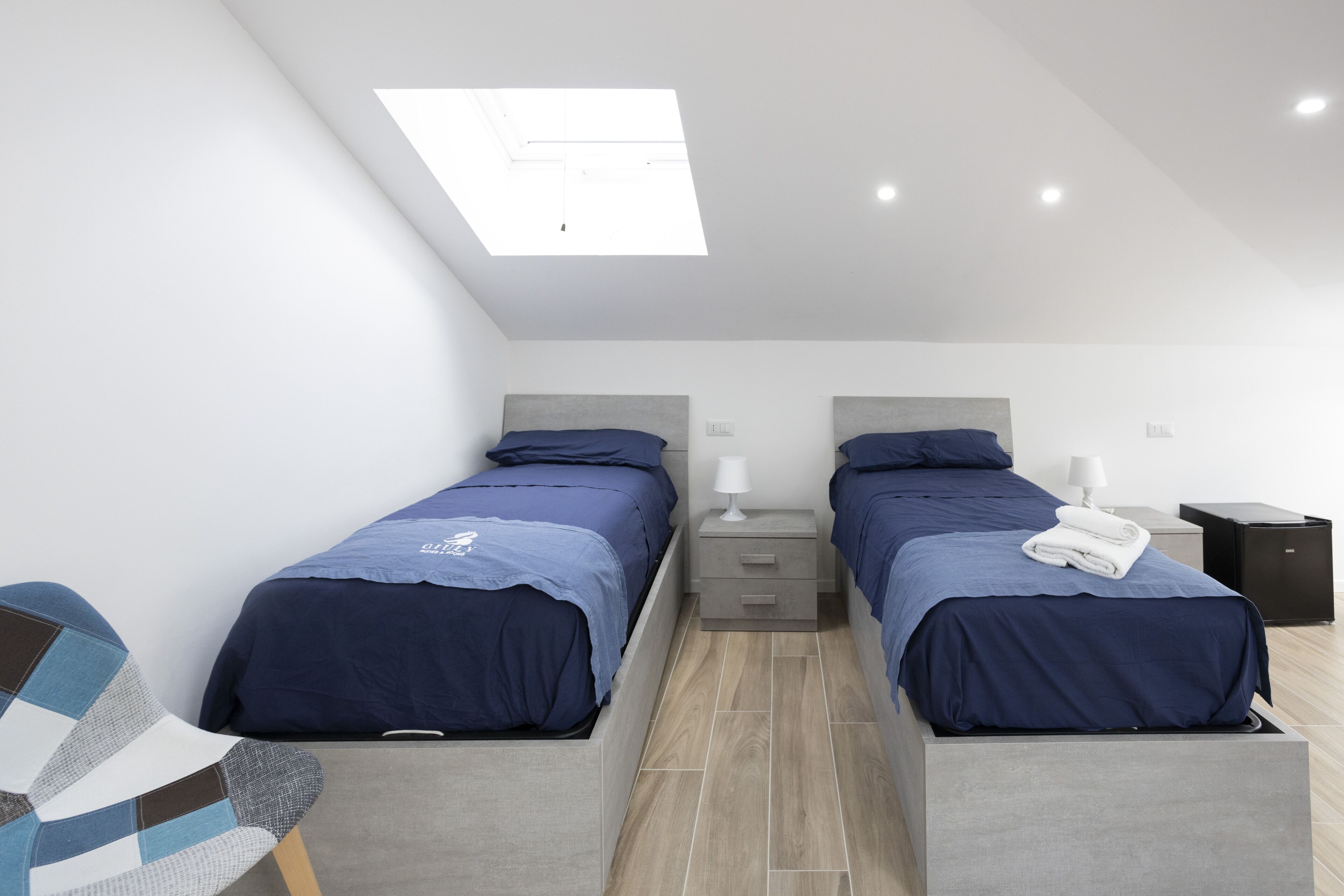 A modern bedroom with two beds, a skylight, and a small bedside table with a lamp.