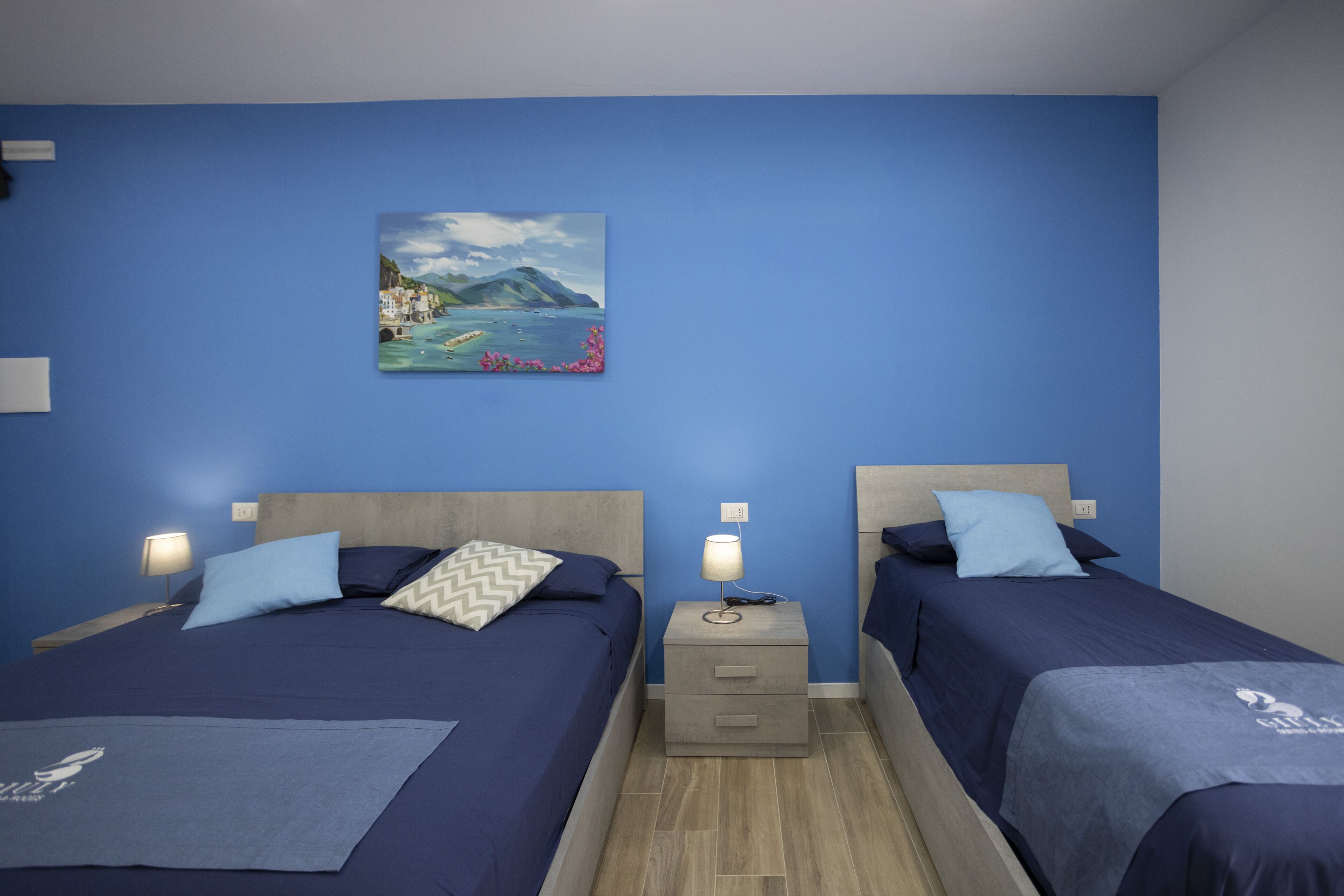 Quarto de hotel com duas camas, uma mesinha de cabeceira com luminária e um quadro na parede.