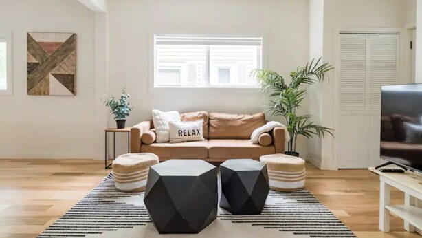 A living room with a beige sofa, two ottomans, a coffee table, a TV, and a window with blinds. - Central District 5BR — Walk to Capitol Hill, Cal Anderson Park & Broadway (Seattle)