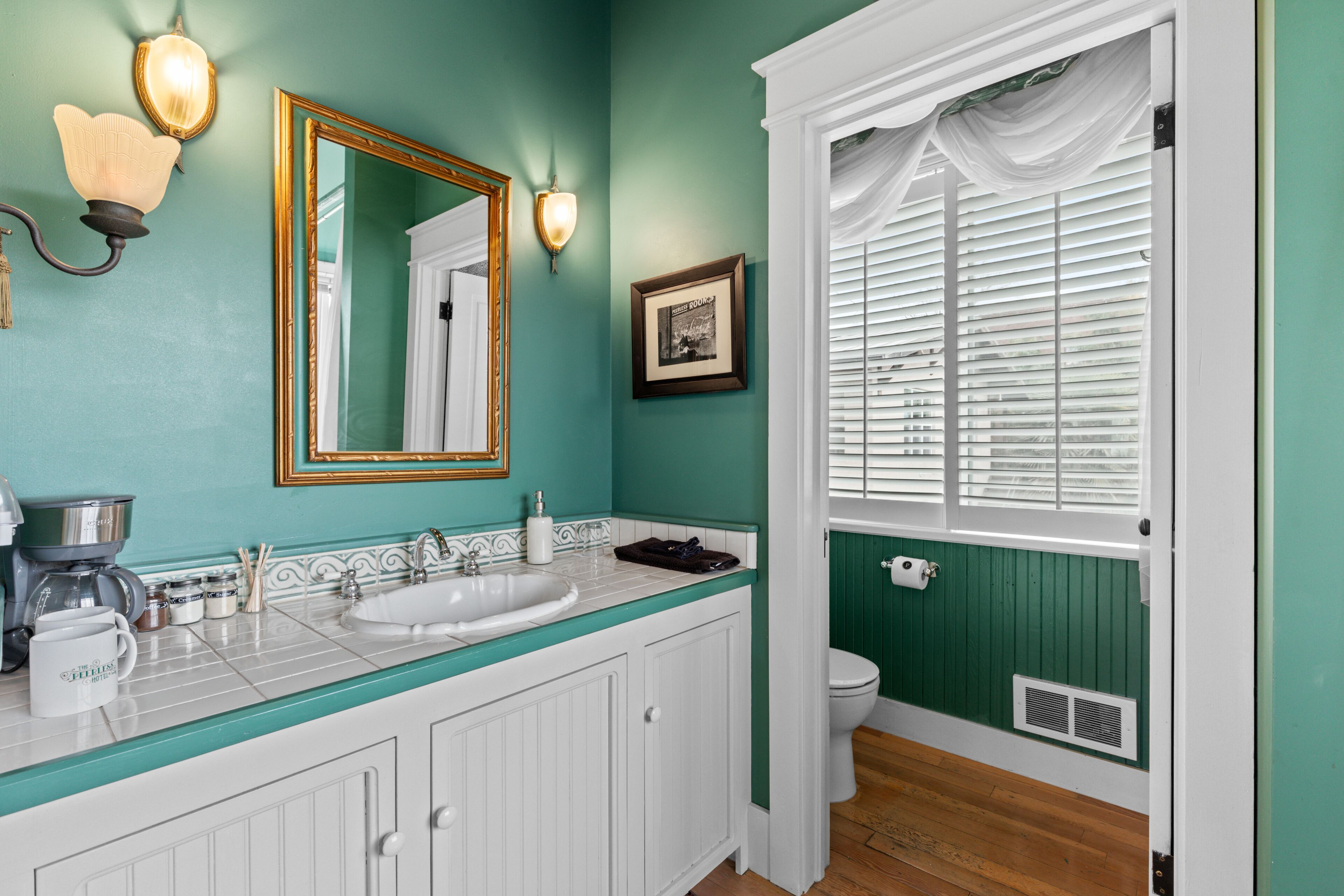 A bathroom with teal walls, white cabinetry, a gold-framed mirror, a sink, and a toilet.
