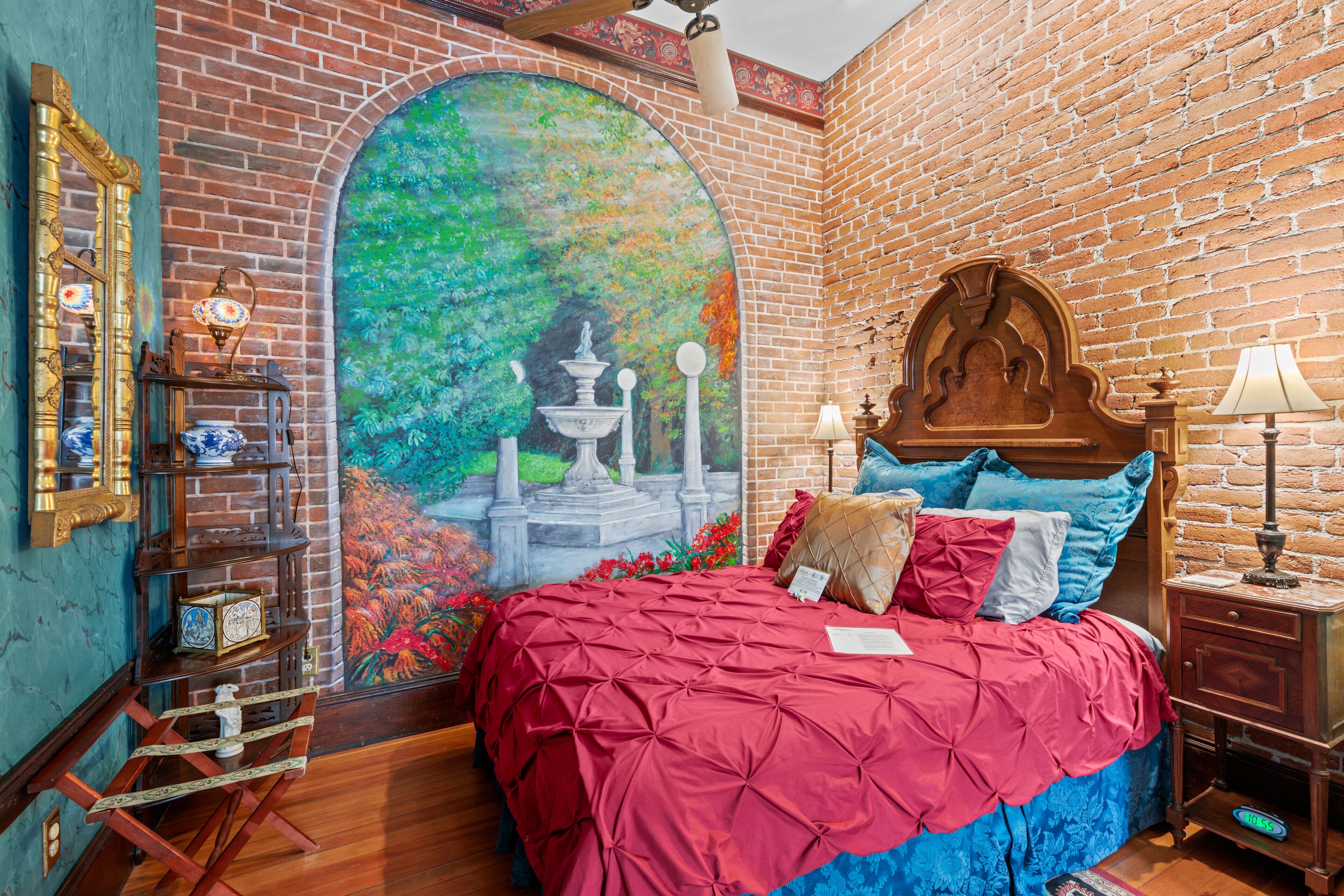 A bedroom with a brick wall, a mural, a bed with red and blue bedding, a wooden headboard, and a nightstand with a lamp.