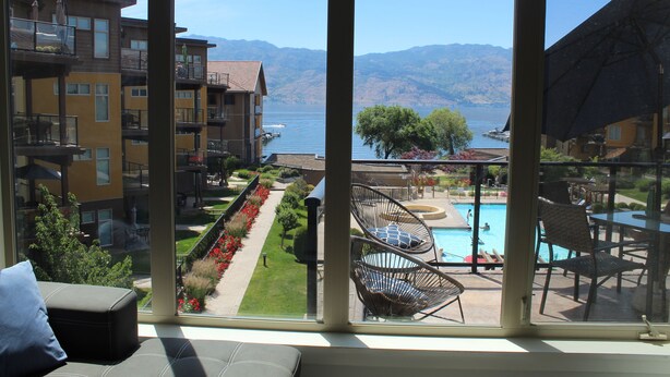 Vista desde una ventana con piscina, zona de estar exterior y montañas al fondo.