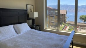 Habitación de hotel con una cama grande, una mesita de noche con lámpara y ventana con vistas a edificios y una masa de agua.