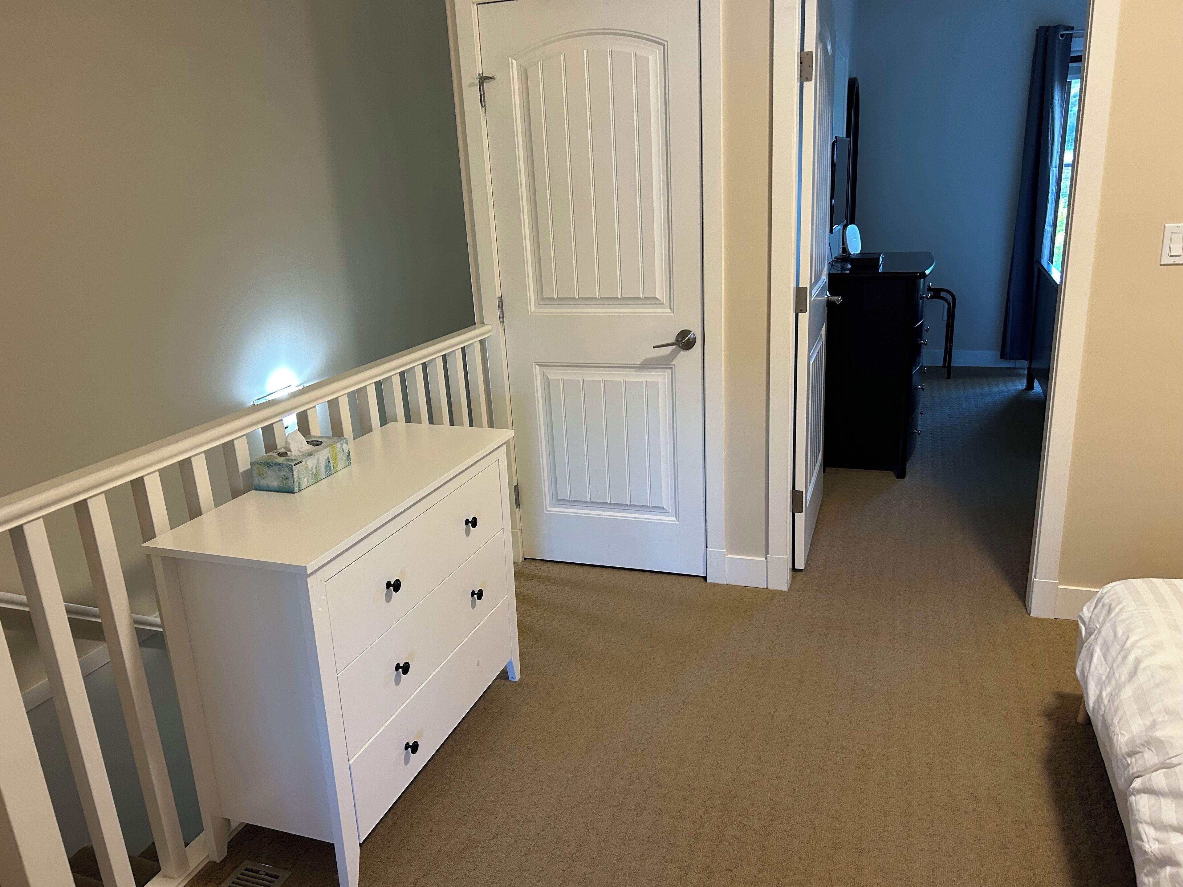 A bedroom with a white dresser, a door, a bed, and a small desk with a mirror.