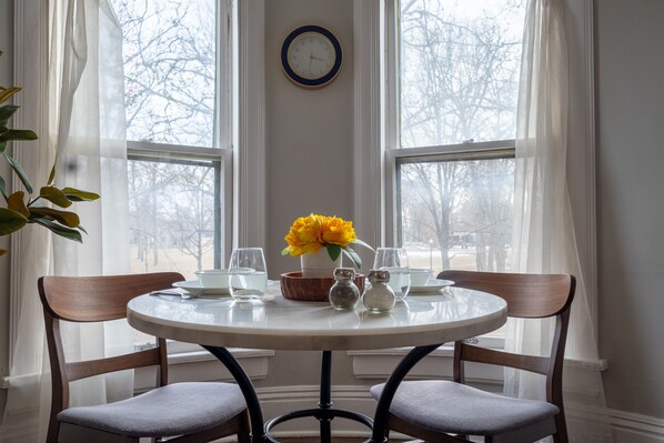 A round dining table with chairs, a clock on the wall, and a window with curtains.