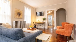 A living room with a sofa, a small table with a tray, an orange chair, a desk with a chair, a TV, and a radiator.