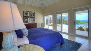 A bedroom with a large bed, a ceiling fan, a nightstand, a lamp, and a view of a swimming pool through glass doors.
