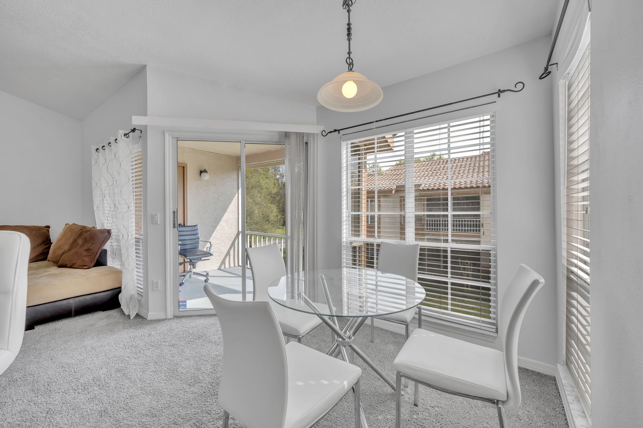 A bright, white interior with a glass table and chairs, a sofa, and a view through a window with blinds.