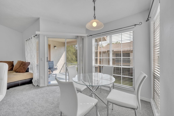 A bright, white interior with a glass table and chairs, a sofa, and a view through a window with blinds.