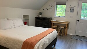 Une chambre à coucher avec un lit, un bureau, une chaise, un seau rouge et une fenêtre donnant sur un paysage verdoyant.