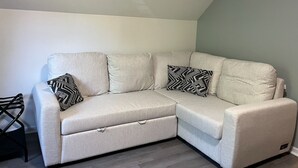 Un canapé d’angle de couleur claire, en forme de L, agrémenté de deux coussins aux motifs noir et blanc, une chaise en métal noir et un sol gris.