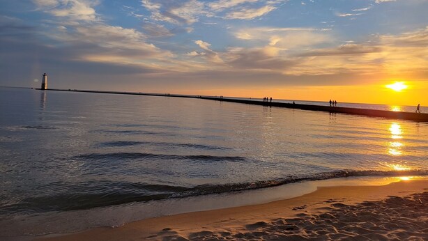 Une plage paisible avec une jetée qui s’avance dans l’eau, un phare à son extrémité, et le soleil se couchant à l’horizon.