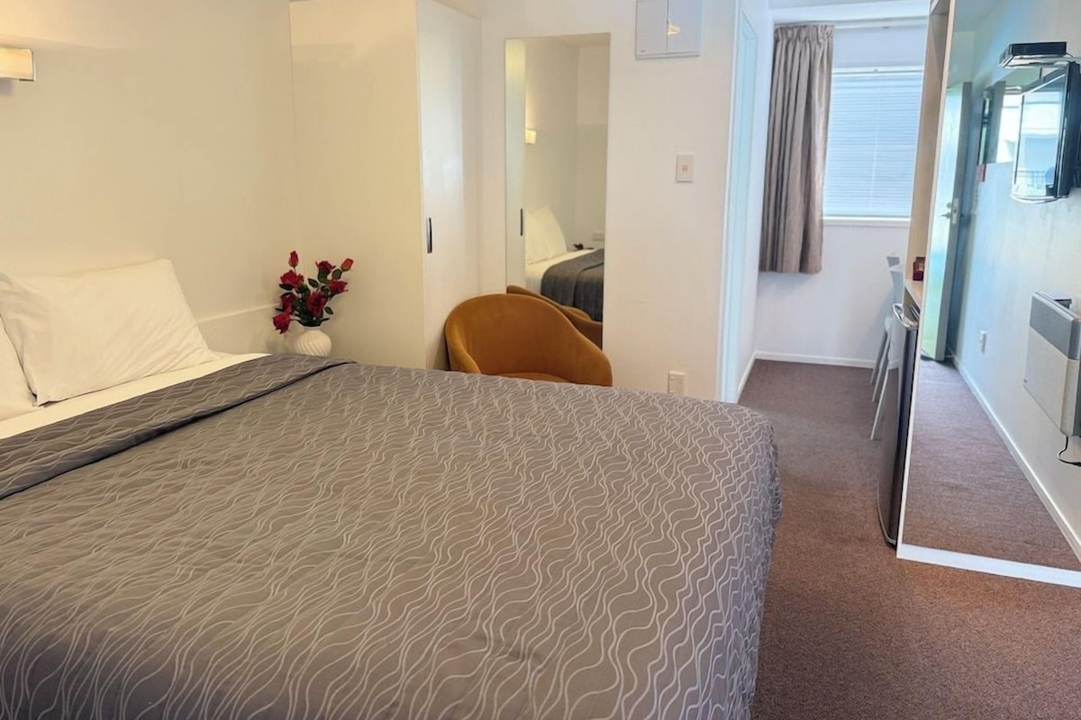 A hotel room with a bed, a chair, a small table with a vase of flowers, a mirror, and a window with curtains.