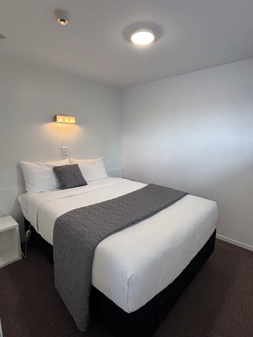 Ein Einzelbett mit weißer Matratze und grauer Decke, ein Nachttisch mit Lampe sowie eine Wandleuchte.