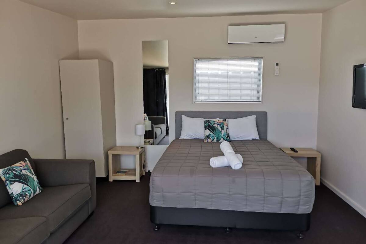 A hotel room with a bed, a sofa, a small table, a mirror, a window with blinds, an air conditioning unit, and a television.