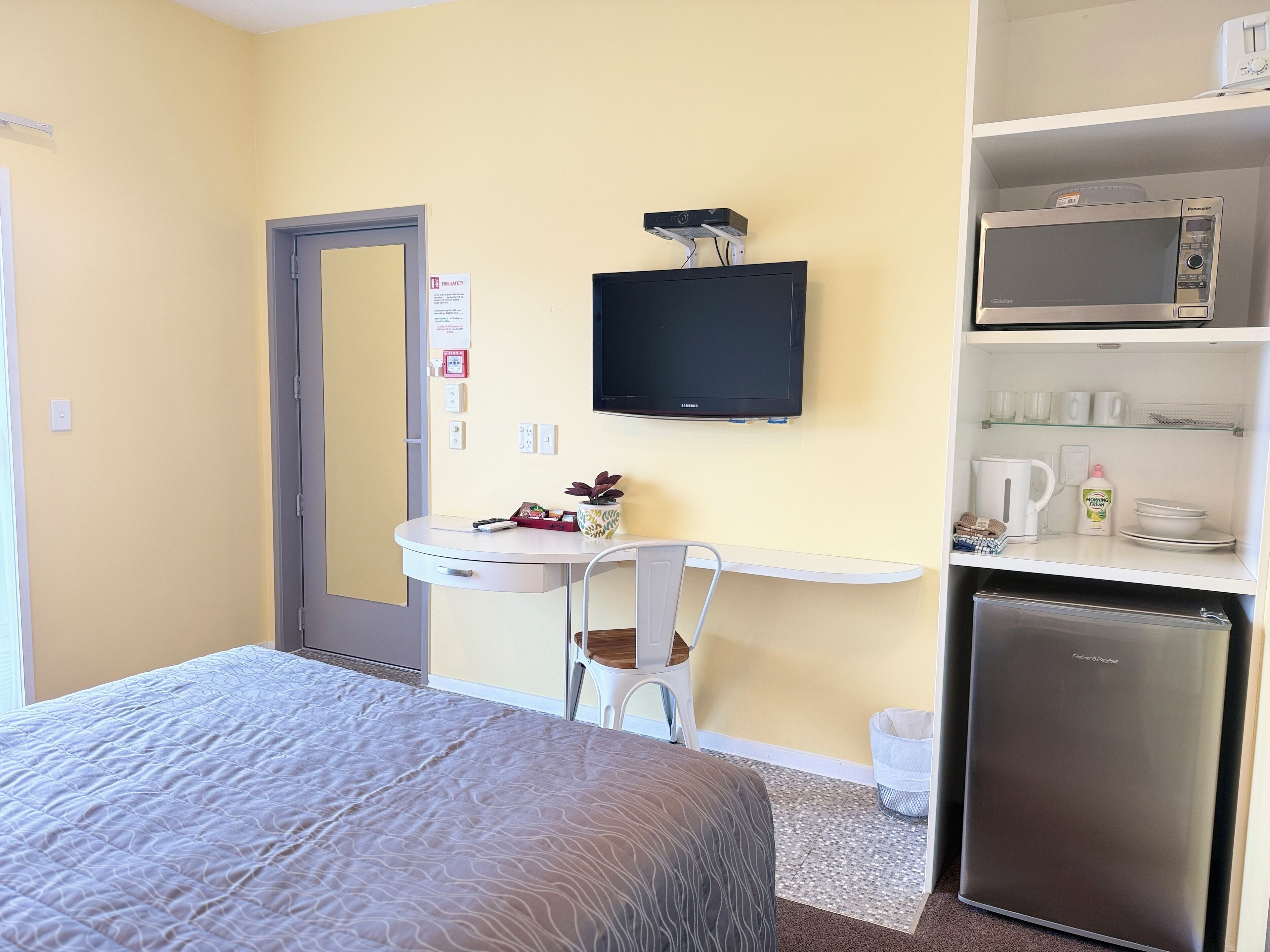 A hotel room with a bed, a desk with a chair, a microwave, a refrigerator, and a small shelf with cups and a kettle.