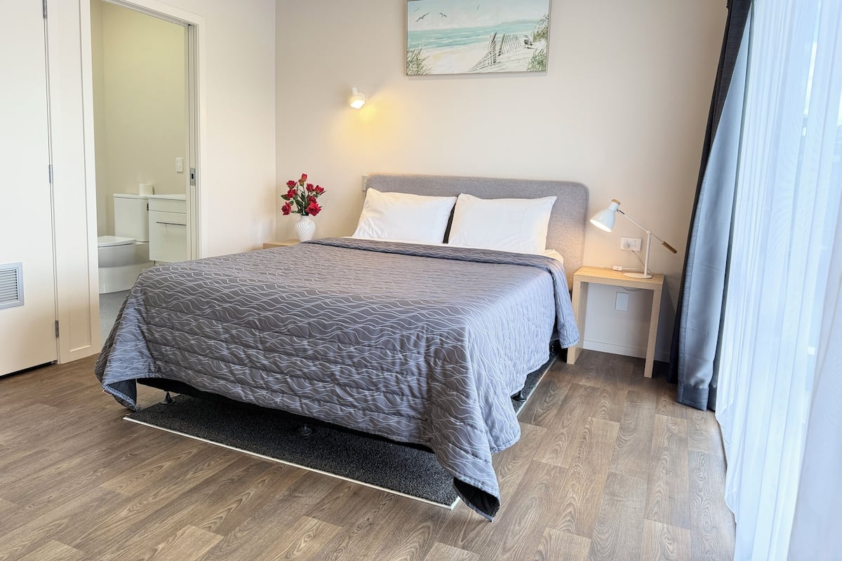 A neatly arranged bedroom with a bed, bedside tables, a nightstand, a wall-mounted lamp, and a painting above the bed.