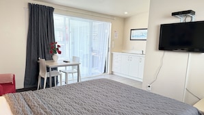 A hotel room with a bed, a desk with a chair, a TV mounted on the wall, and a view of the kitchen through the sliding glass door. - Morrinsville Motel (Morrinsville)