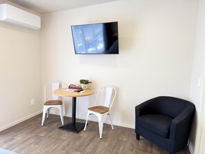 A room with a round table, two chairs, a black armchair, a flat-screen TV mounted on the wall, and an air conditioning unit. - Morrinsville Motel (Morrinsville)