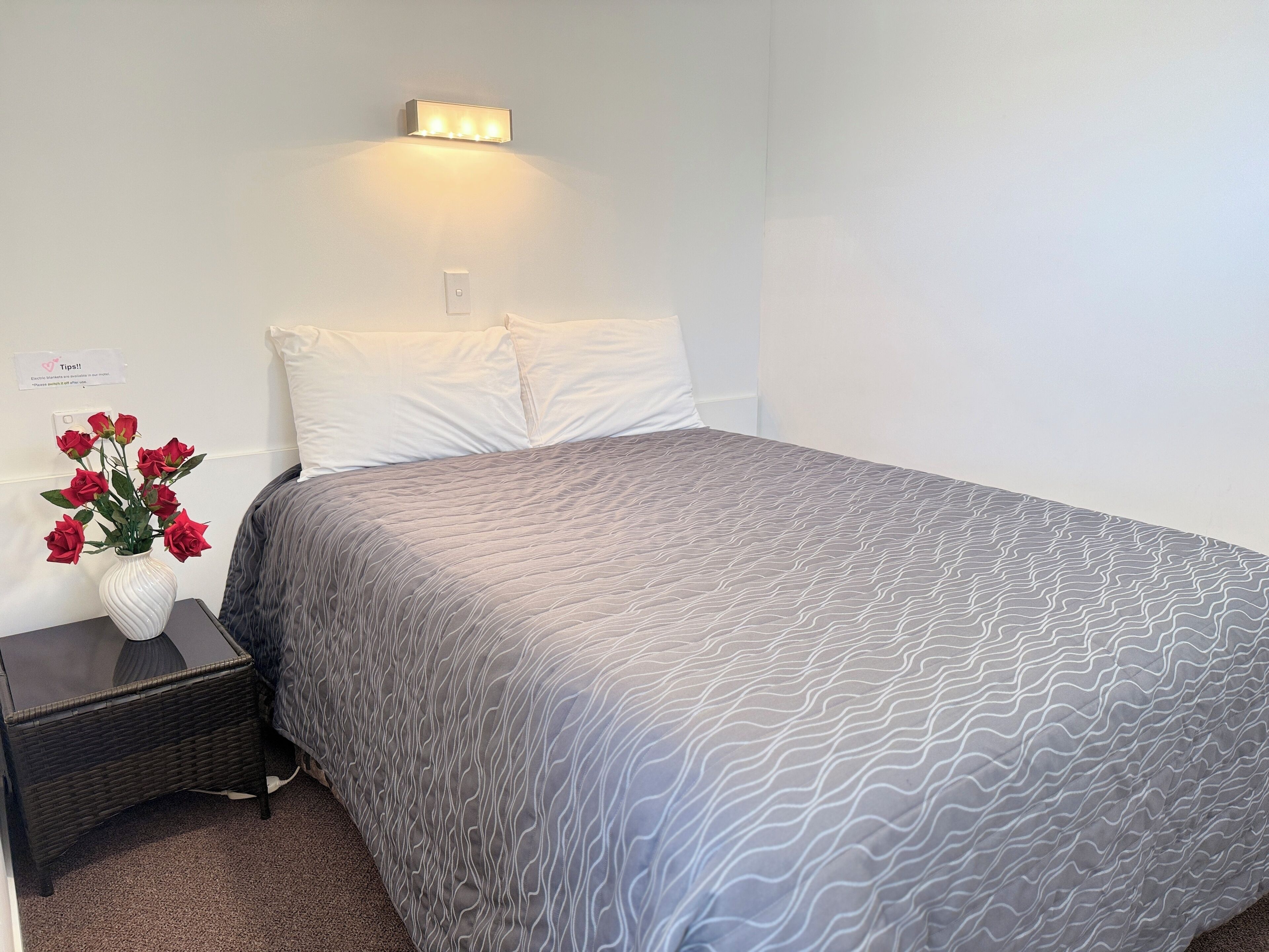 A neatly made bed with a patterned blanket, a white vase with red flowers on a bedside table, and a wall-mounted light fixture.