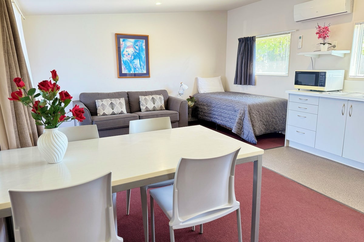 A modern hotel room with a dining area, a sofa set, a bed, a small kitchenette, and a painting on the wall.