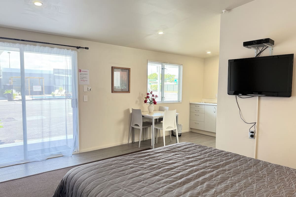 A hotel room with a bed, a desk with chairs, a television mounted on the wall, and a window with blinds.
