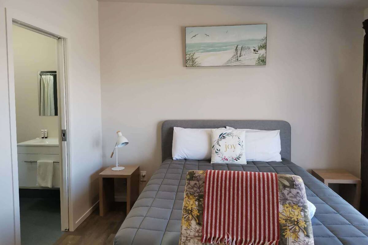 A bedroom with a bed, a nightstand, a lamp, a painting, and a bathroom visible through an open door.