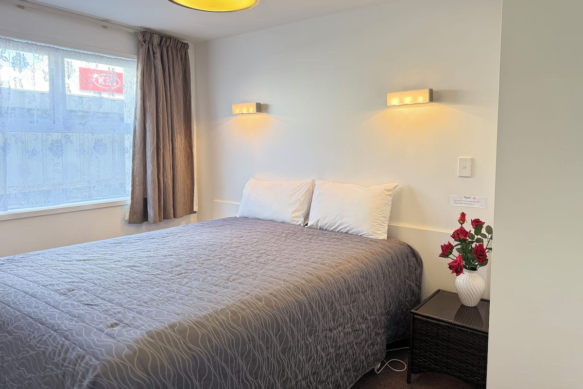 A hotel room with a bed, a nightstand, a vase with flowers, and a window with curtains.