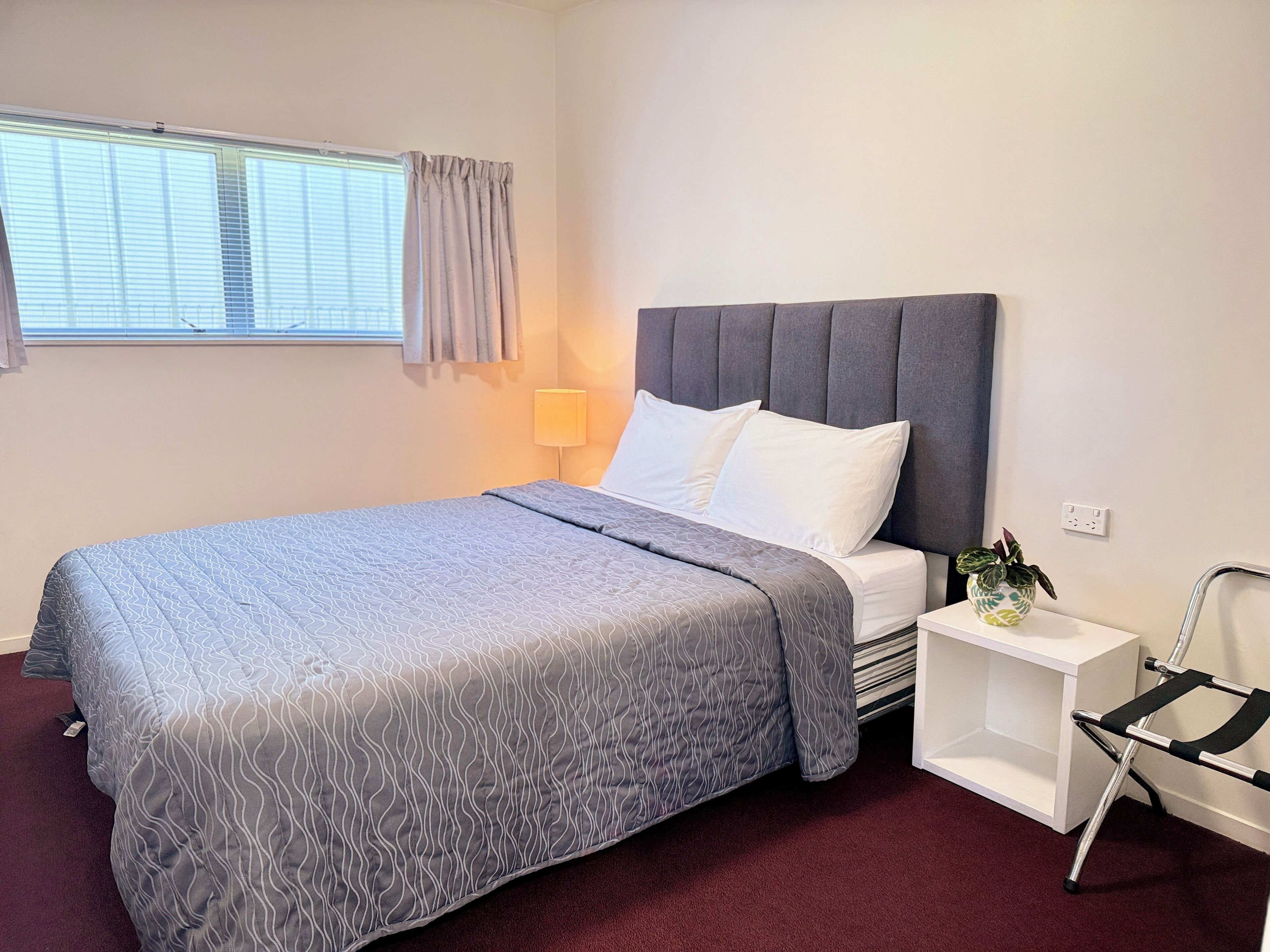 A neatly made bed with a grey patterned bedspread and white pillows, a bedside table with a lamp, a small plant, and a metal chair.