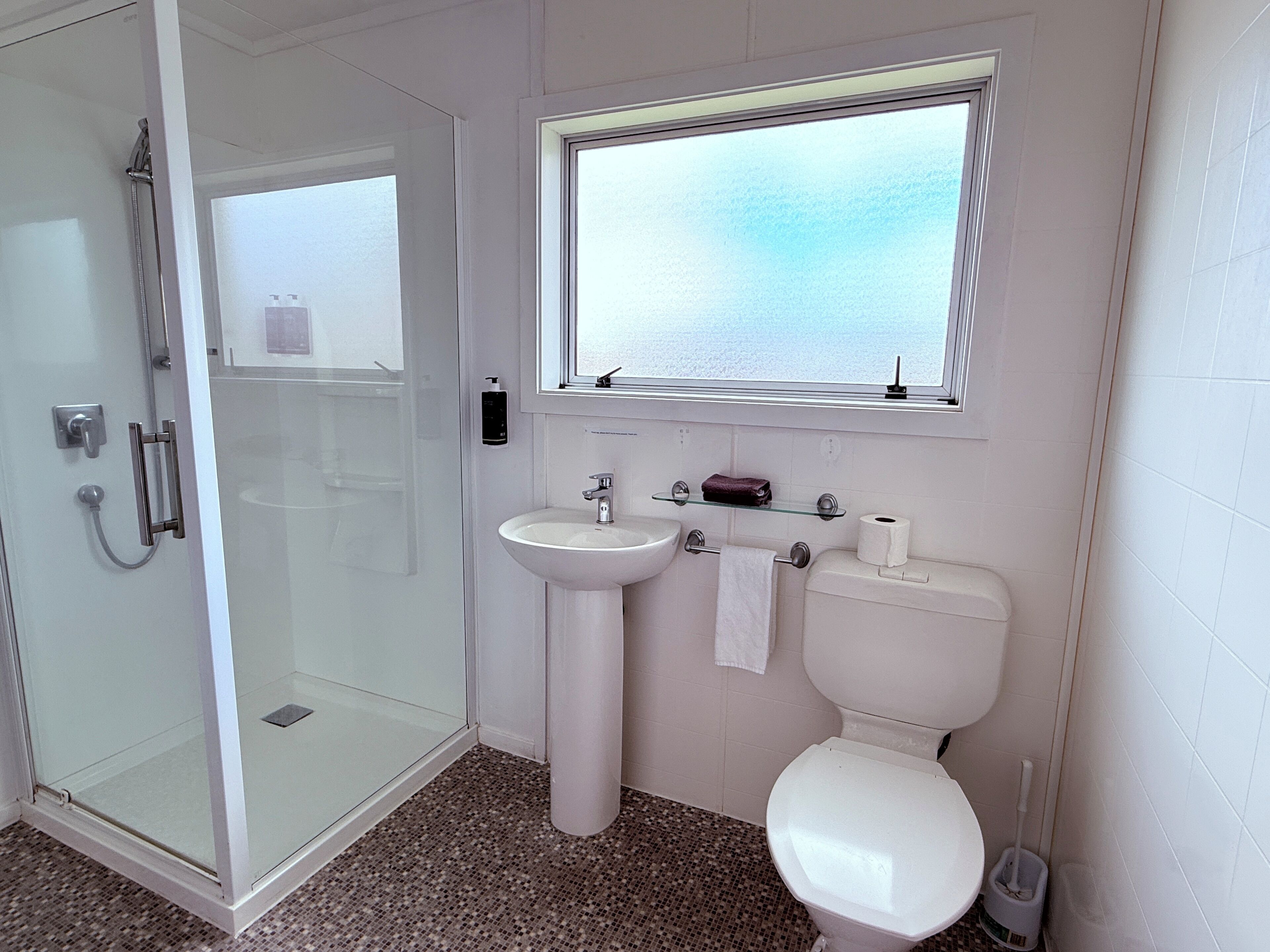 Een badkamer met een douchecabine van glas, een wastafel op voet, een toilet en een raam boven de wastafel.