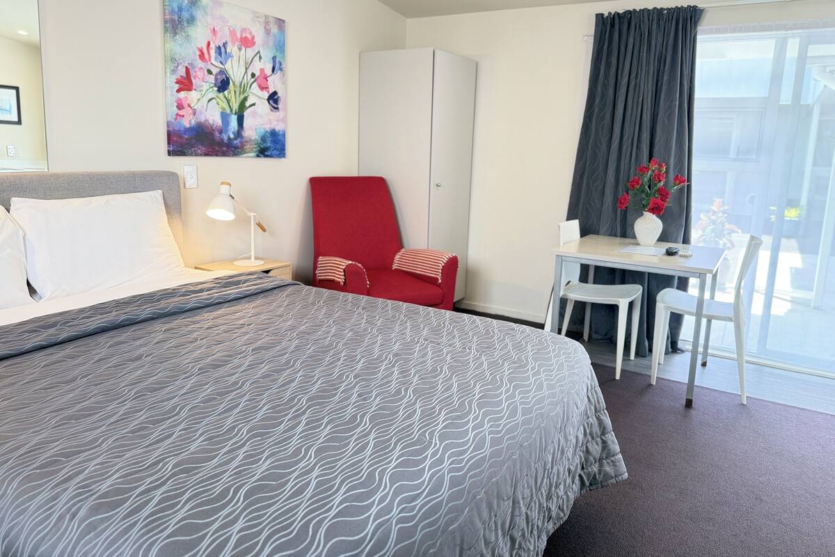 A hotel room with a bed, a desk, a chair, a red armchair, a painting, and a view of the outdoors.
