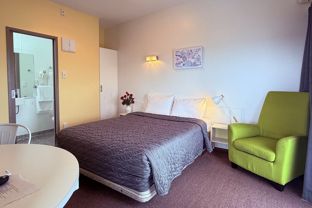 A hotel room with a bed, a green armchair, a small table, and a painting on the wall.