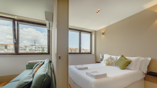A modern bedroom with a large bed, a sofa, and a view of the city through large windows. - Legacy Oporto Design Apartment G by Vacationy (Porto)
