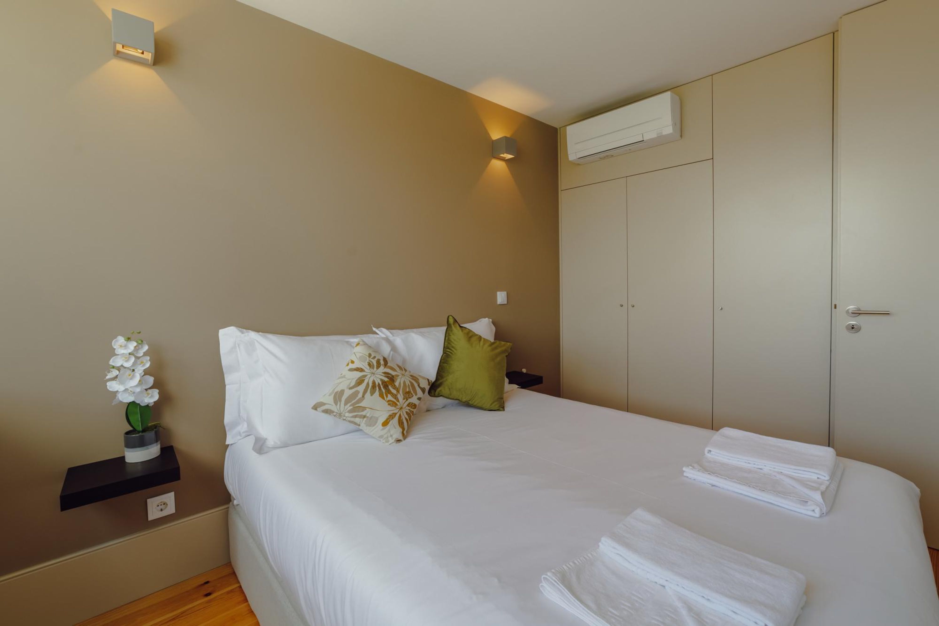 A neatly made bed with white linens, a small nightstand with a vase of white flowers, and a wall-mounted light fixture.