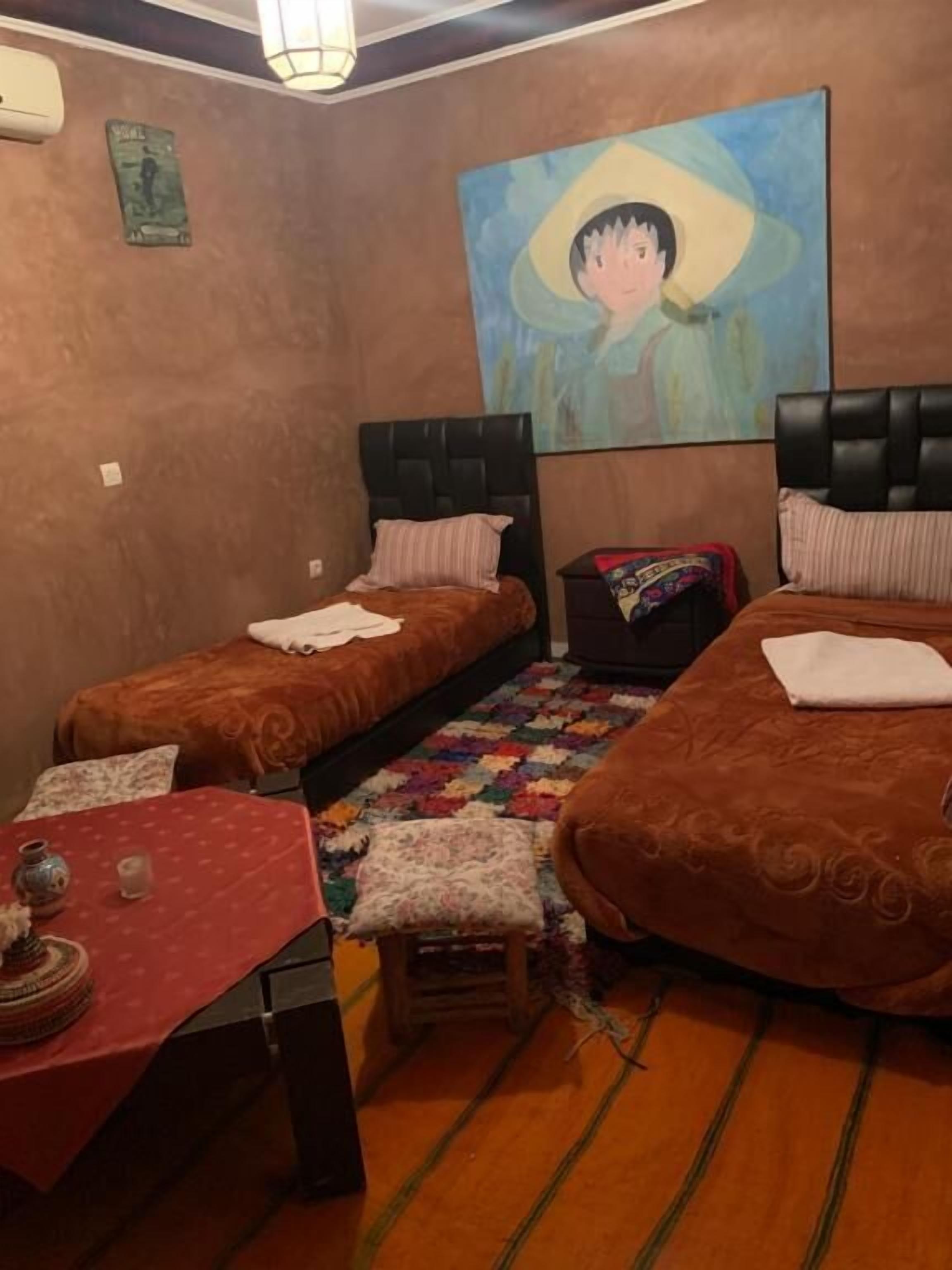A room with two single beds, a small table with a red tablecloth, and a quilted rug.