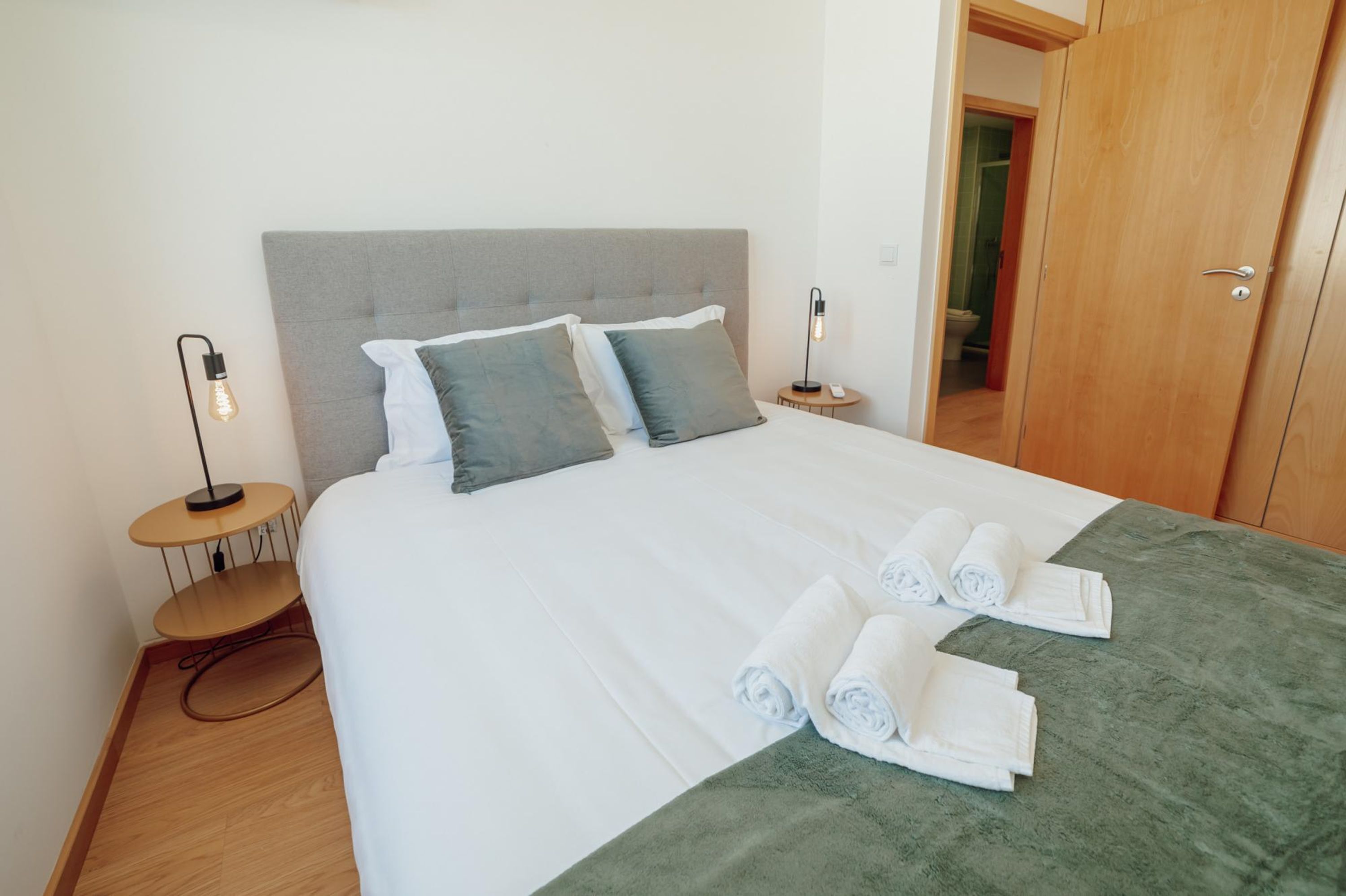 Un lit bien fait, avec du linge de lit blanc et gris, un plaid vert et des serviettes pliées posées sur le lit.