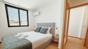 Ein modernes Schlafzimmer mit einem großen Bett, einem grauen Kopfteil, einem Nachttisch mit Lampe und einem Fenster mit Blick auf Gebäude.