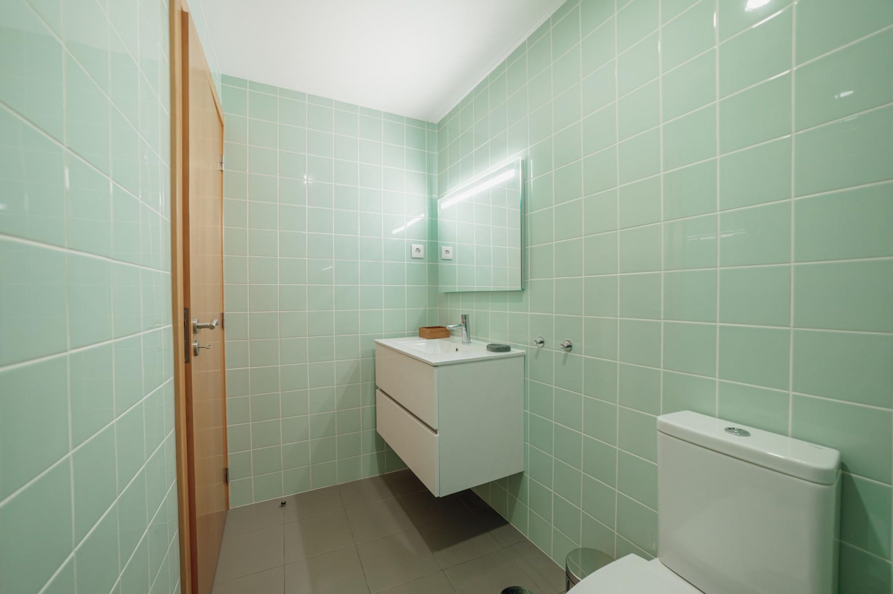 A bathroom with green tiles, a white sink, a mirror, and a toilet.