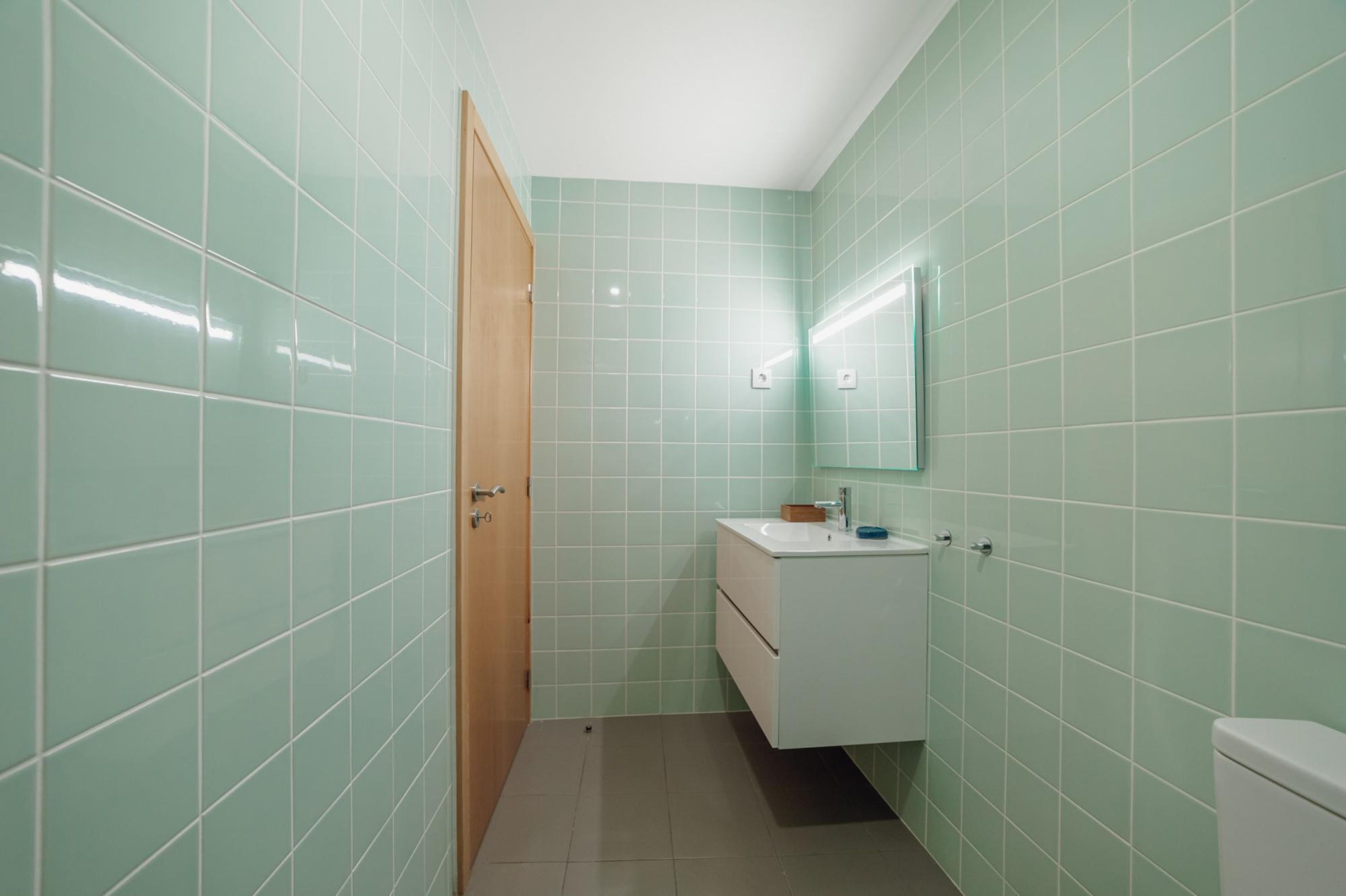 Casa de banho com azulejos verdes, porta de madeira, lavatório branco e espelho.