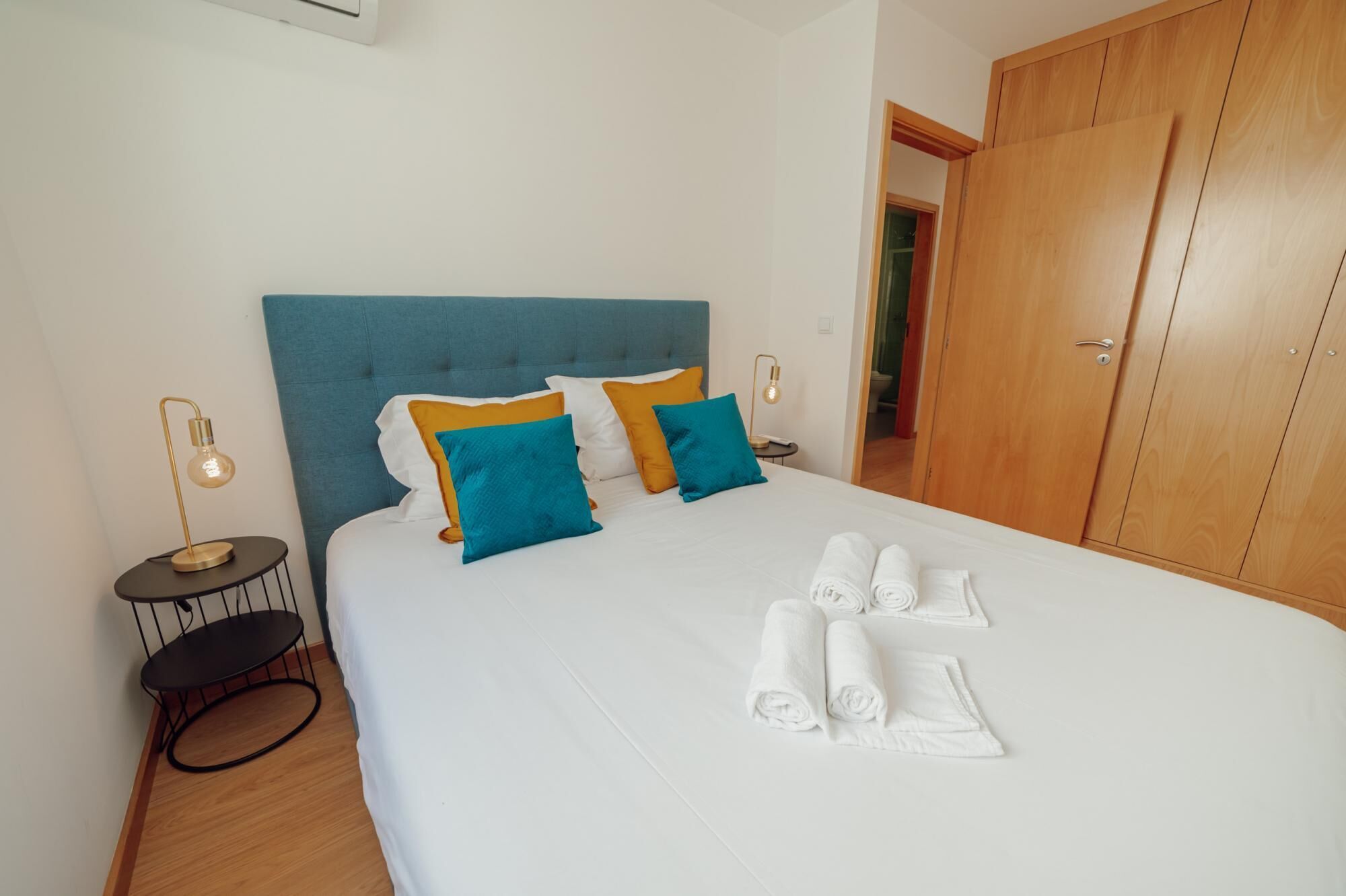 A neatly made bed with white linens, a blue headboard, and several pillows in blue and yellow. There are two bedside tables with lamps, and a wooden wardrobe is visible in the background.